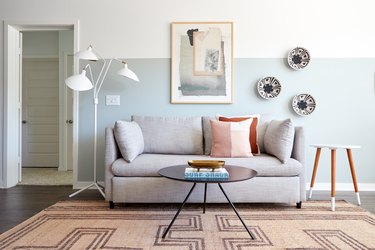 Eclectic living room with gray couch, light blue wall, white floor lamp, round coffee table, side table, art, pillows, rug, plates on wall.